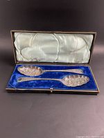 Two ornate Victorian berry spoons resting inside their original hinged presentation case with blue velvet interior and white satin lid lining, viewed from above