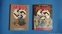 Both volumes of MAUS by Art Spiegelman placed side by side on blue surface showing front covers and condition.