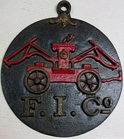 Front view showing round cast iron plaque with antique fire pumper painted red on black background, with "F. I. Co" text below.