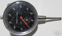 Front view of dial indicator gauge showing black dial face with numeric graduations and red needle pointers.