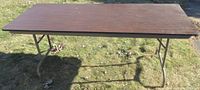Full top view of six-foot rectangular folding table with dark wood grain fiberboard surface outdoors on grass.