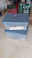Two stacked clear plastic totes with gray lids containing Christmas lights, unopened.