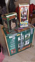 Photo of three boxed holiday items: Christmas tree stand, musical animated Santa figure in box, electrified church decoration in box
