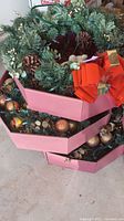 Three artificial Christmas wreaths stacked in pink boxes. Each wreath has integrated green wire lights, pinecones, white berries. Orange bows and small plush bear decorations visible on two wreaths.