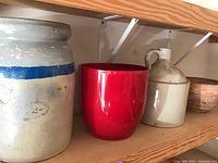 Shelved view showing multiple pottery items including the two gallon crock with blue stripe, red ceramic flower pot, beige pottery jug, and smaller clay flower pot.