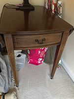 Close-up of wooden end table showing main surface, one drawer with metal handle, and tapered leg with decorative inlay.