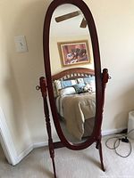 Full view of the standing oval mirror in a bedroom setting, showing wooden frame and reflective surface, angled slightly to the left.