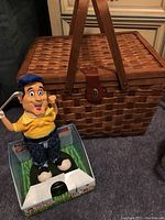 Photo showing woven wooden picnic basket with dual handles and leather strap, alongside a colorful dancing golfer toy in original packaging.