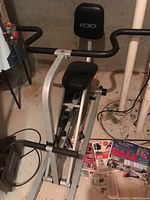 Front angled view of manual rowing machine showing padded seat, backrest and handlebars in basement setting.