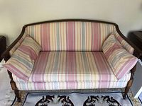 Front view of the small settee sofa showing wooden frame with ornate arms, nail head trim, and pastel striped upholstery with two side cushions.
