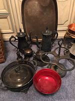 Overview of all silver plated items including coffee pots, serving tray, covered casserole dish, cream and sugar set, small bowl, red glass bowl, and condiment holder, all heavily tarnished.