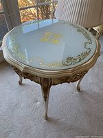 View of the glass top showing ornate gold detailing and round shape, placed near a window and chair.