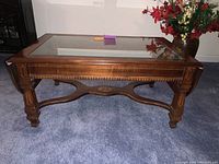 Front-side view of wooden coffee table with glass inset top, decorative legs and cross stretcher, standing on blue carpet.