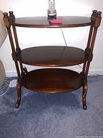 Front view of the wooden table showing three oval shelves and claw feet on a carpeted floor with remote on top.
