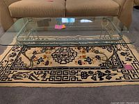 Rectangular glass top table with silver and gold metal ornate base placed on a decorative black and white rug with floral patterns beneath a beige sofa.