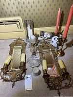 Two ornate brass wall mirrors with candle holders and cream faux candles placed on carpet with other candlesticks and small glassware.