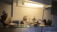 Overview of lot on glass shelf showing roosters, pottery, floral plaque, brass items, clock, and irons