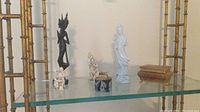 Group of Asian themed figures displayed on glass shelf including a tall black wooden figurine, a light blue ceramic figure, an ivory ceramic two-children figurine, a black and white small item, and marble lighter cases.