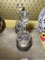 Two Hawkes cut glass decanters placed inside ornate silver plate holders on carpet with relaxed lighting showing the overall presentation and scale.
