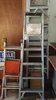 Two 6-foot aluminum ladders leaning in garage space. Visible signs of paint splatter, dirt marks, and a metal work tray with circular cutouts on top of one ladder. Included an orange folding chair in foreground.