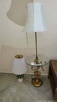 Two table lamps placed side by side: one tall lamp with white pleated shade and brass round glass table surface, and one shorter ceramic lamp with detailed green fruit-shaped base and white pleated shade.