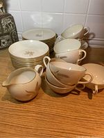 Photo showing 11 bread plates stacked, 9 cups, 6 saucers stacked and 1 creamer with the wheat motif and gold trim on a wooden surface