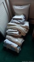 View of stacked assorted linens including folded blankets, sheets, and a large pillow against a wall.