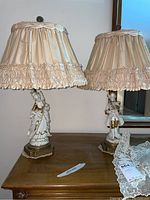 Pair of vintage porcelain lamps with figurine bases topped with pink pleated satin shades with ruffles, sitting on wooden table.