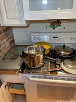 Photo of assortment of pots and pans on stove including stainless steel pot, black frying pan with glass lid, yellow frying pan, small skillet, and cooking racks
