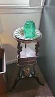 Full view of antique wooden plant stand displaying its sculpted legs and drawer, green crackle glass vase on top, and copper tea kettle placed underneath.