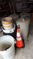 Photo of multiple gray plastic trash cans with lids, orange traffic cone, plastic baskets, and white buckets in a storage area
