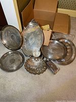 Photo showing seven vintage silver plated serving pieces arranged on carpet near an open cardboard box, varying shapes and designs, showing surface tarnish and use wear.