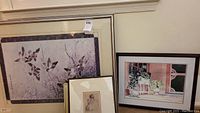 Full view of three framed artworks leaning against the wall: a large Asian bird print with blue border, a smaller framed black and white drawing, and a framed watercolor window scene.
