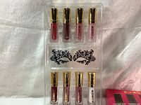 Top view of 8 mini BUXSOM lip gloss bottles with gold caps in a plastic tray insert showing various shimmery shades.