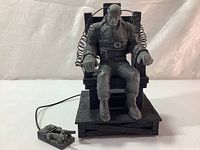 Front view of Marv figurine seated in electric chair with remote control device in front