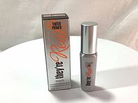 Photo of the Benefit They're Real! tinted eyelash primer bottle and box on a white round surface, showing front of box and bottle labeling.