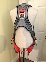 Photo showing the back of the full body safety harness on a mannequin, detailing the construction, stitching, shoulder strap design, and metal D-ring attachment points.