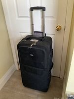 Front view of the black rolling camera gear case with retractable handle extended, standing upright against door.