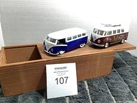 Two Volkswagen bus model vehicles placed in an open wooden box, side view.