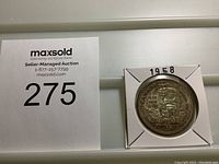 Photo of the reverse side of a 1958 Canadian silver dollar coin showing Indigenous totem design, the dates 1858-1958 and text 'Canada', 'British Columbia', and 'Dollar'. Coin is secured in a white cardboard holder with the year 1958 written on it.