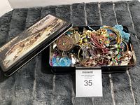 Image showing the open decorative tin box containing a disorganized assortment of costume jewelry including bracelets, bangles, necklaces and pins.
