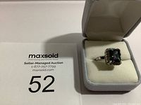 Close-up of ring in white cushioned box, showing the front view with square mystic topaz and halo stones.