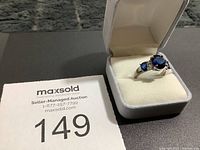 Photo of ring in white cushioned ring box showing blue gemstones, silver band, and auction lot number 149.