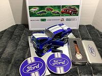 Photo showing blue and white 40th Anniversary Ford Mustang model with open doors, two round purple Ford coasters, and the Ford watch in its tin box.
