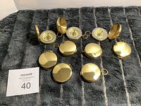Nine brass pocket compasses arranged on a gray plush surface. Five compasses show closed covers, four compasses have their brass covers open revealing the compass dials with directional markings and keyring loops attached.