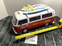 Side view of red and white Volkswagen bus toy with measuring tape showing approximate length around 12 inches, surfboards on roof, and lot number card visible.