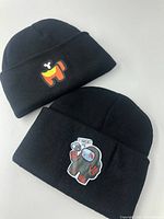 Two new black knit beanie hats laid flat, showing front cartoon patches.