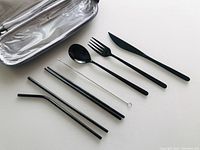 Matte black travel utensils arranged next to silver insulated carrying case showing knife, fork, spoon, chopsticks, two straws, and cleaning brush.