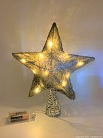 Lit silver wire star-shaped Christmas tree topper with warm white LED lights glowing, coiled base and AAA batteries beside it for scale.
