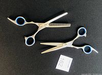 Two pairs of silver-colored hair scissors with blue rubber finger holes on a black background, showing one thinning scissors and one straight scissors.
